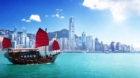 A traditional red-sailed junk boat gliding across Victoria Harbour in Hong Kong, with the city's iconic skyline and towering skyscrapers illuminated in the background.