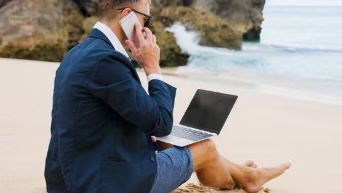 Man sitting on the beach while working on a laptop demonstrating how technology for business travel is all about making things simple